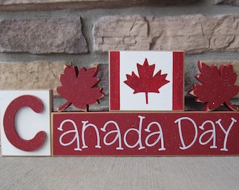 Może przedstawiać: Drewniany znak z napisem "Canada Day" białymi literami na czerwonym tle. Znak jest ozdobiony czerwonymi liśćmi klonu i flagą Kanady.