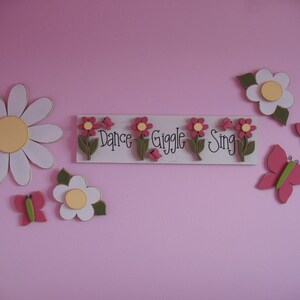 May include: A pink wall with a white wooden sign that says "Dance Giggle Sing" with pink flowers and green leaves. There are also white and pink flowers and butterflies on the wall.