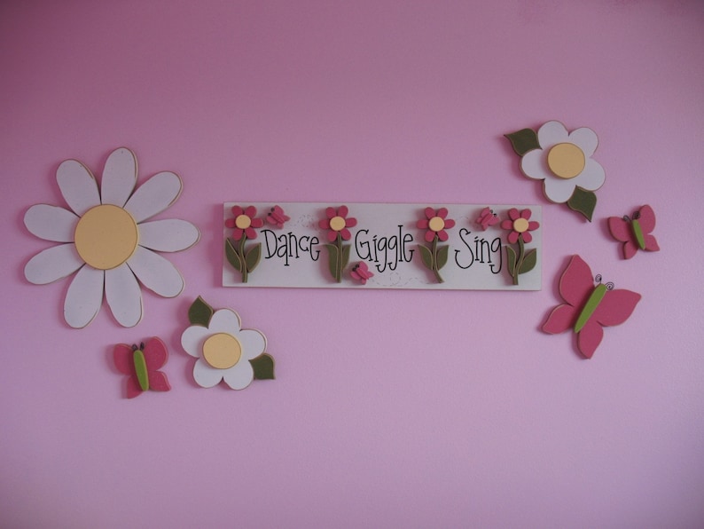 May include: A pink wall with a white wooden sign that says "Dance Giggle Sing" with pink flowers and green leaves. There are also white and pink flowers and butterflies on the wall.