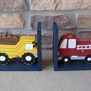 May include: A pair of wooden bookends shaped like a yellow dump truck and a red fire truck. The bookends are painted with details and have a blue base.