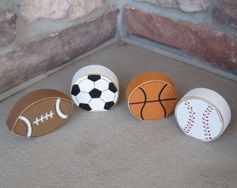 May include: Four wooden ball ornaments painted to look like different sports: a football, soccer ball, basketball, and baseball. The ornaments are all white with the sports painted on them in their respective colors.