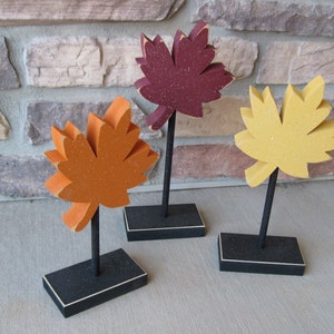 May include: Three wooden fall leaves on black stands. The leaves are painted in shades of orange, yellow, and red.