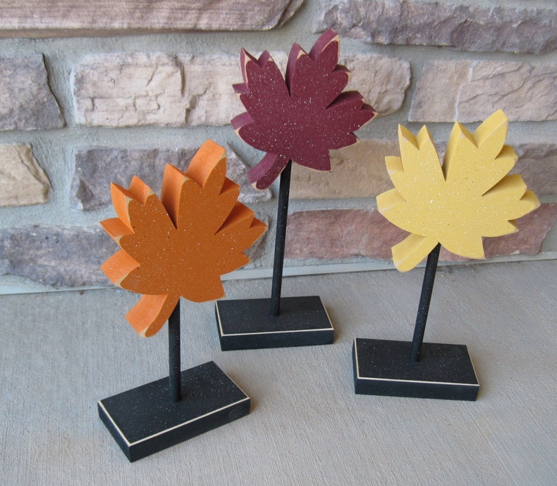 May include: Three wooden fall leaves on black stands. The leaves are painted in shades of orange, yellow, and red.