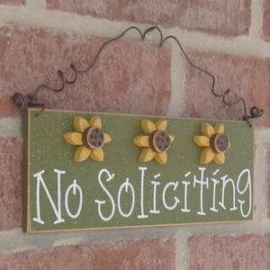 May include: A green wooden sign with the words "No Soliciting" in white lettering. The sign is decorated with three yellow sunflowers with brown centers.