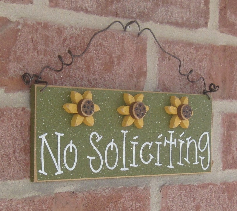 May include: A green wooden sign with the words "No Soliciting" in white lettering. The sign is decorated with three yellow sunflowers with brown centers.