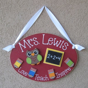 May include: Red oval wooden sign with the text "Mrs. Lewis" in white. The sign has a brown owl with green wings, a yellow chalkboard with the equation "2+2=4", and four colorful pencils with the words "Love", "Teach", "Inspire" written around them.