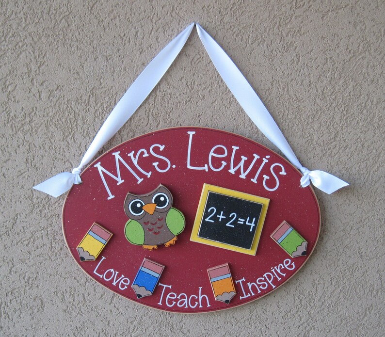 May include: Red oval wooden sign with the text "Mrs. Lewis" in white. The sign has a brown owl with green wings, a yellow chalkboard with the equation "2+2=4", and four colorful pencils with the words "Love", "Teach", "Inspire" written around them.