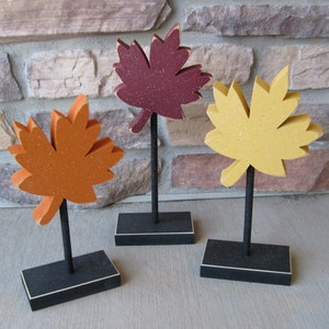 May include: Three wooden fall leaf decorations on black stands. The leaves are painted in shades of orange, yellow, and maroon.