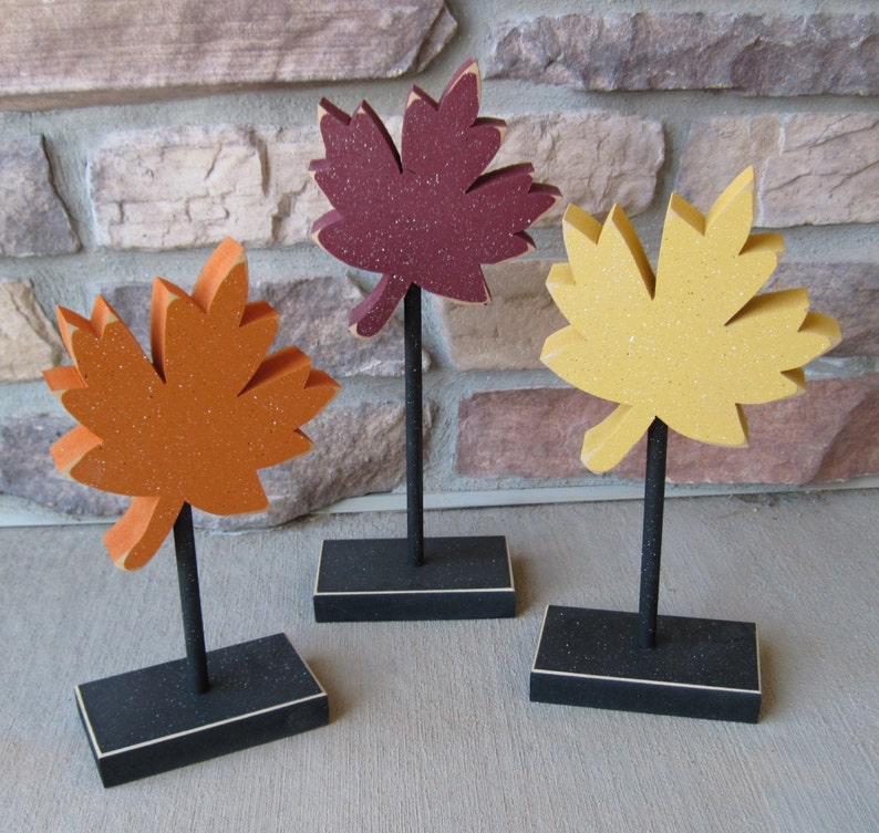 May include: Three wooden fall leaf decorations on black stands. The leaves are painted in shades of orange, yellow, and maroon.