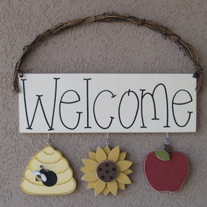 May include: A white wooden sign with the word "Welcome" written in black. The sign has three hanging decorations: a yellow beehive with a black bee, a yellow sunflower, and a red apple.