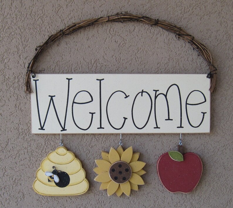 May include: A white wooden sign with the word "Welcome" written in black. The sign has three hanging decorations: a yellow beehive with a black bee, a yellow sunflower, and a red apple.