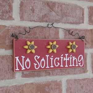 May include: Red wooden sign with white text that reads "No Soliciting". The sign is decorated with three yellow sunflowers with brown centers. The sign is hanging from a black wire.