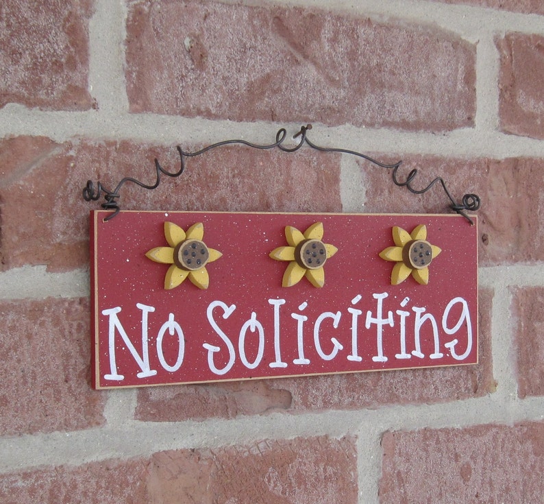 May include: Red wooden sign with white text that reads "No Soliciting". The sign is decorated with three yellow sunflowers with brown centers. The sign is hanging from a black wire.