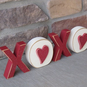 May include: Wooden XOXO letters with a red glitter finish and gold trim. The O's feature a white background with a red heart. The letters are arranged on a gray surface, perfect for Valentine's Day or a romantic gift.