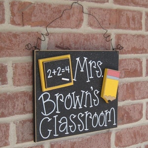 May include: Black chalkboard sign with white lettering that reads "Mrs. Brown's Classroom". The sign has a yellow and black chalkboard with the equation "2+2=4" written on it and a yellow pencil with a red eraser.