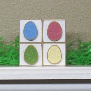 May include: Four wooden blocks with a white background and a painted egg design on each block. The eggs are blue, pink, green, and yellow. Each egg has a glittery texture.
