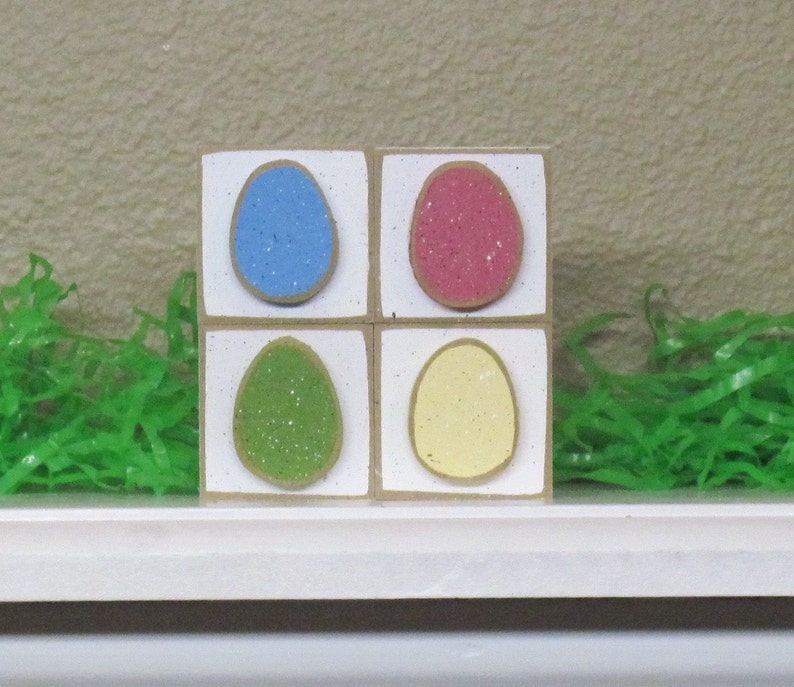 May include: Four wooden blocks with a white background and a painted egg design on each block. The eggs are blue, pink, green, and yellow. Each egg has a glittery texture.