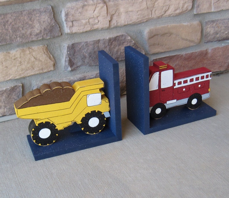 May include: A pair of wooden bookends featuring a yellow dump truck and a red fire truck. The bookends are painted with details and have a blue base.