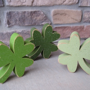 May include: Three wooden shamrock decorations in different shades of green. The shamrocks are painted with a sparkly finish.