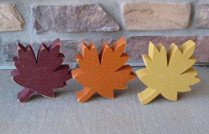 K&ouml;nnte beinhalten: Drei h&ouml;lzerne Ahornbl&auml;tter in Herbstfarben. Die Bl&auml;tter sind burgunderrot, orange und gelb.