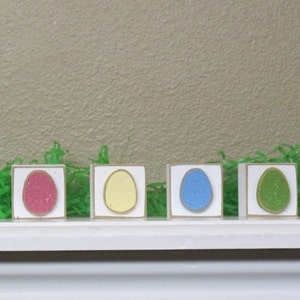 May include: Four wooden blocks, each painted white with a different colored egg shape inside. The eggs are pink, yellow, blue, and green.