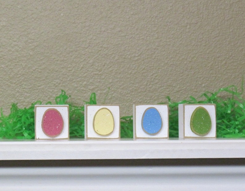 May include: Four wooden blocks, each painted white with a different colored egg shape inside. The eggs are pink, yellow, blue, and green.