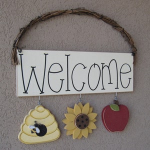 May include: A white wooden sign with the word "Welcome" written in black. The sign is hanging from a brown vine wreath. There are three wooden decorations hanging from the sign: a beehive with a bee, a sunflower, and a red apple.