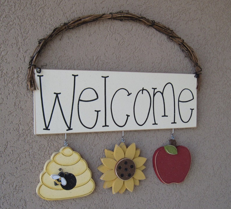 May include: A white wooden sign with the word "Welcome" written in black. The sign is hanging from a brown vine wreath. There are three wooden decorations hanging from the sign: a beehive with a bee, a sunflower, and a red apple.