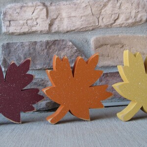 K&ouml;nnte beinhalten: Drei h&ouml;lzerne Herbstbl&auml;tter in verschiedenen Farben. Die Bl&auml;tter sind burgunderrot, orange und gelb. Sie sind alle mit Glitzerlack bemalt.