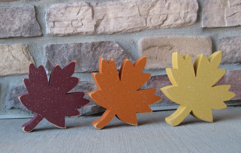 K&ouml;nnte beinhalten: Drei h&ouml;lzerne Herbstbl&auml;tter in verschiedenen Farben. Die Bl&auml;tter sind burgunderrot, orange und gelb. Sie sind alle mit Glitzerlack bemalt.