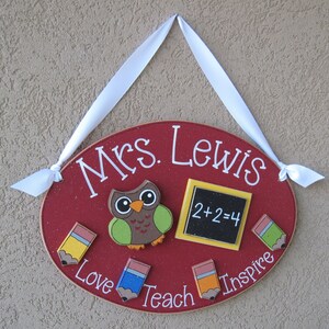 May include: Red oval wooden sign with the text "Mrs. Lewis" in white. The sign has a brown owl with green wings, a yellow chalkboard with the equation "2+2=4", and four colorful pencils with the words "Love", "Teach", "Inspire" written around them.
