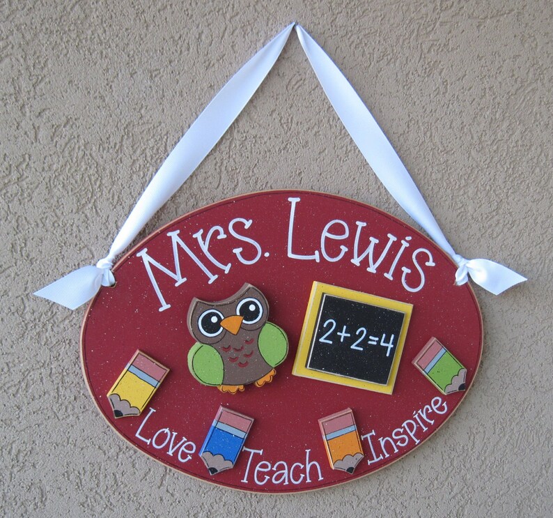 May include: Red oval wooden sign with the text "Mrs. Lewis" in white. The sign has a brown owl with green wings, a yellow chalkboard with the equation "2+2=4", and four colorful pencils with the words "Love", "Teach", "Inspire" written around them.