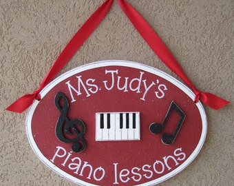 May include: Oval-shaped sign with the text "Ms. Judy's Piano lessons" in white lettering. The sign is red with a white border and features a treble clef, a piano keyboard, and a musical note. A red ribbon is attached for hanging.