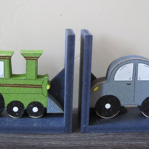 May include: A pair of bookends featuring a green toy train and a gray toy car. The train has black wheels and red stripes. The car has black wheels and a yellow headlight. The bookends are made of wood and have a blue base.