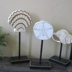 May include: Three decorative wooden figurines on black stands. The figurines are a seashell, a sand dollar, and a conch shell. The seashell is white with gold accents, the sand dollar is white with a black starfish design, and the conch shell is white with brown accents.