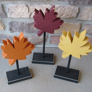 May include: Three wooden fall leaf decorations on black stands. The leaves are red, orange, and yellow with a speckled texture.