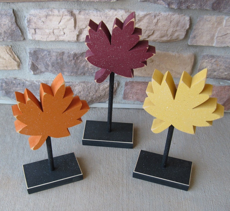 May include: Three wooden fall leaf decorations on black stands. The leaves are red, orange, and yellow with a speckled texture.
