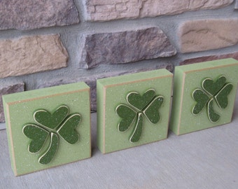 May include: Three green wooden blocks with a shamrock design. Each block has a green shamrock with a white outline on a green background.