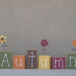 May include: Wooden blocks spelling out the word "AUTUMN" with a sunflower, a leaf, and a pumpkin on top of each block.