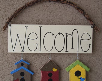 May include: A wooden welcome sign with three birdhouses hanging below. The birdhouses are painted in different colors: blue, red, and yellow. Each birdhouse has a small bird perched on the roof.
