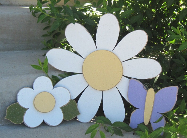 May include: Three wooden decorations: a large white daisy with a yellow centre, a smaller white daisy with a yellow centre, and a purple butterfly with a yellow body.
