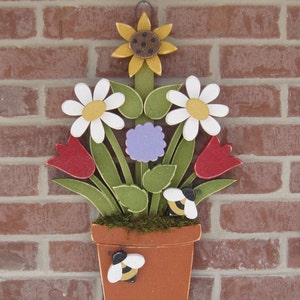 May include: Wooden flower pot with a sunflower, daisies, tulips, and bees. The pot is painted brown and has a green moss-like texture around the top.