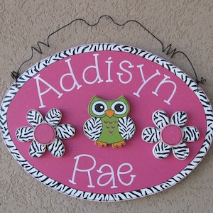 May include: Pink oval wooden sign with a black and white zebra print border. The sign has the name "Addisyn Rae" written in white paint. There is a green owl with black eyes and a pink flower with a zebra print on either side of the name.