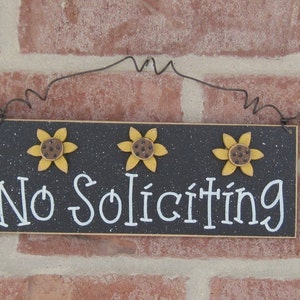 May include: Black wooden sign with white text that reads "No Soliciting". The sign is decorated with three yellow sunflowers with brown centers.