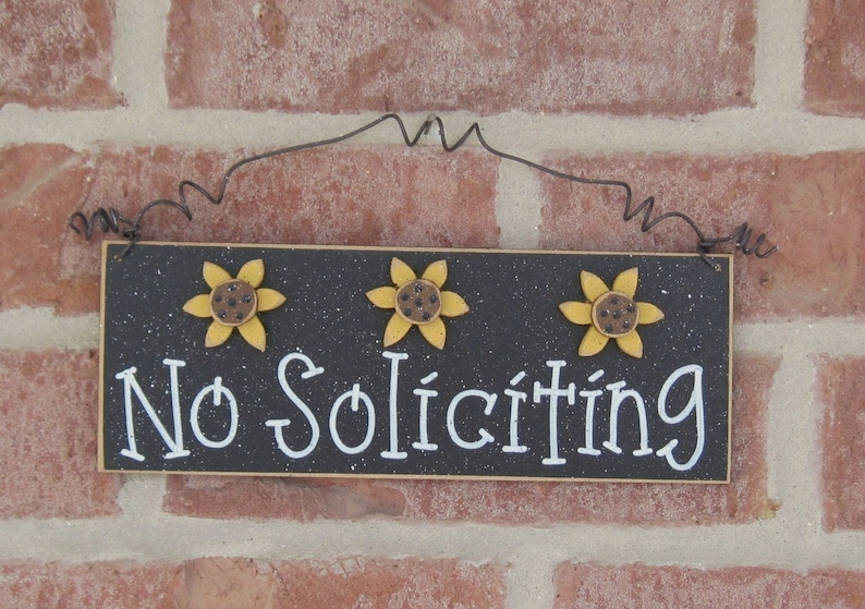 May include: Black wooden sign with white text that reads "No Soliciting". The sign is decorated with three yellow sunflowers with brown centers.