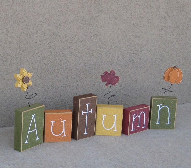 May include: Wooden blocks spelling out the word "autumn" with a sunflower, leaf, and pumpkin on top of each block.