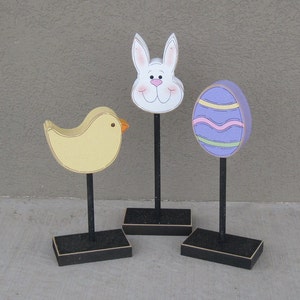 May include: Three wooden Easter decorations on black stands. A yellow chick, a purple and white striped egg, and a white bunny with a pink nose.