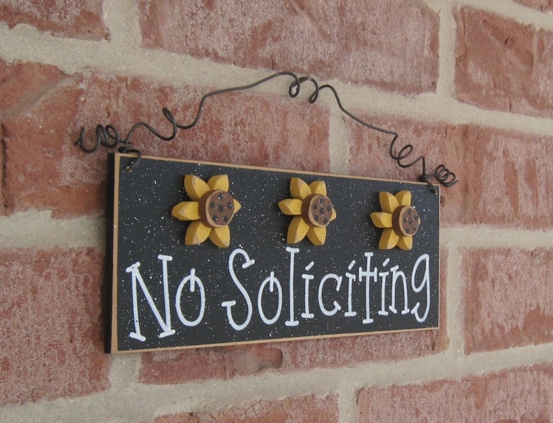 May include: Black wooden sign with white text that reads "No Soliciting". The sign is decorated with three yellow sunflowers with brown centers. The sign is hanging from a black wire.