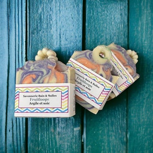 May include: Handmade soaps with a marbled effect in purple and orange, each topped with a decorative piece. The soaps are labelled "Savonnerie Bain & Bulles Fruitloops Argile et soie". They are displayed on a teal-painted wooden surface.
