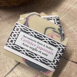 Puede incluir: Una pastilla de jab&oacute;n rectangular con una base de color crema que presenta remolinos de color rosa y gris oscuro. Una etiqueta de papel blanco envuelve el centro, impresa con el texto Savonnerie Bain & Bulles Cerisier japonais Avec argile et soie.
